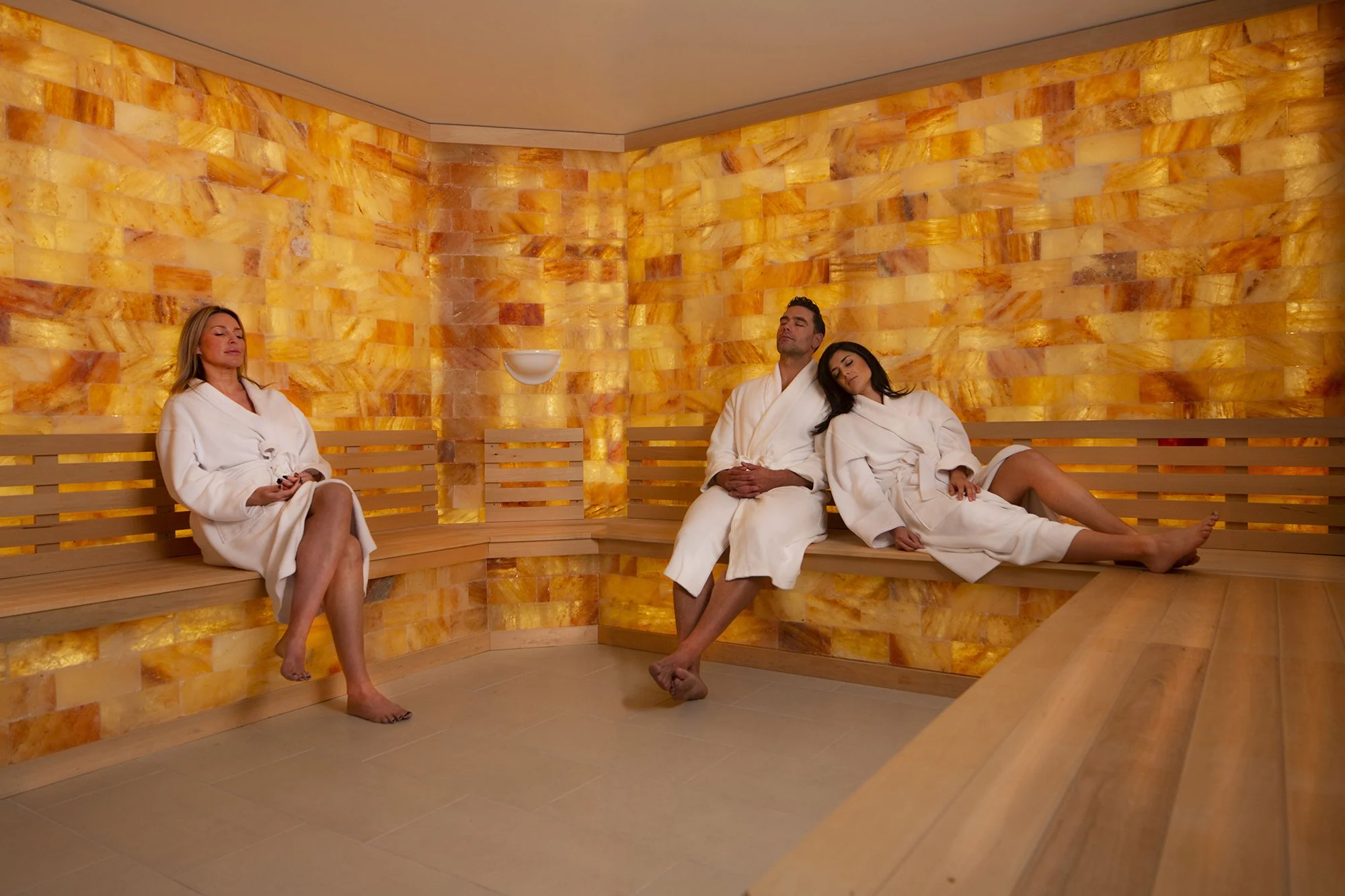 a man and women wrapped in bath towels, relax in a tranquil Himalayan salt room with illuminated salt walls, enjoying the warm glow and therapeutic atmosphere.